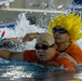 Two individuals are in a swimming pool, both wearing orange shirts and swim goggles. The person in the background is wearing a distinctive bright yellow spiky wig resembling an anime character (likely inspired by Dragon Ball Z's Super Saiyan style). The foreground person wears a beige swim cap, and both appear to be participating in an assisted or recreational swimming activity.