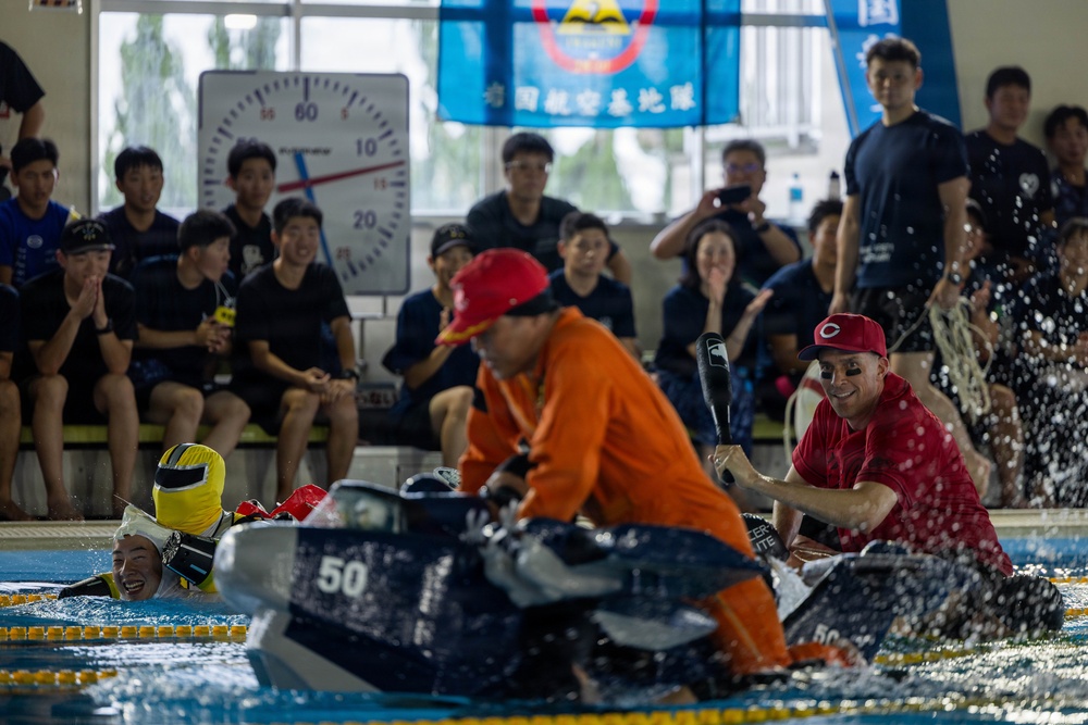 A lively pool event is taking place at an indoor swimming facility, where participants in colorful costumes are competing in what appears to be a cardboard boat race or similar novelty water competition. Two participants in red caps and orange/red shirts are paddling makeshift boats numbered '50' while a person in a yellow costume is in the water laughing. A large crowd of spectators, many wearing dark uniforms, watches enthusiastically from the poolside.