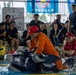 A lively pool event is taking place at an indoor swimming facility, where participants in colorful costumes are competing in what appears to be a cardboard boat race or similar novelty water competition. Two participants in red caps and orange/red shirts are paddling makeshift boats numbered '50' while a person in a yellow costume is in the water laughing. A large crowd of spectators, many wearing dark uniforms, watches enthusiastically from the poolside.