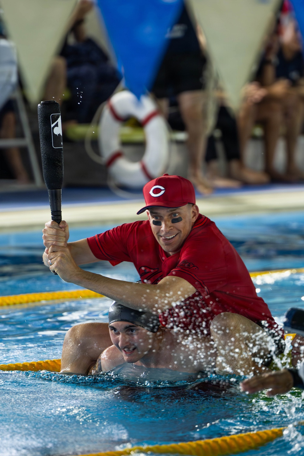 A man wearing a red Cincinnati Reds baseball cap and red shirt, with black eye-black markings under his eyes, is riding on the back of a swimmer in an indoor pool while holding a foam baseball bat aloft. The scene appears to be a fun, competitive pool game or relay event, likely at a recreational or charity event. Lane ropes are visible in the pool, and spectators can be seen in the blurred background.