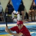 A man wearing a red Cincinnati Reds baseball cap and red shirt, with black eye-black markings under his eyes, is riding on the back of a swimmer in an indoor pool while holding a foam baseball bat aloft. The scene appears to be a fun, competitive pool game or relay event, likely at a recreational or charity event. Lane ropes are visible in the pool, and spectators can be seen in the blurred background.