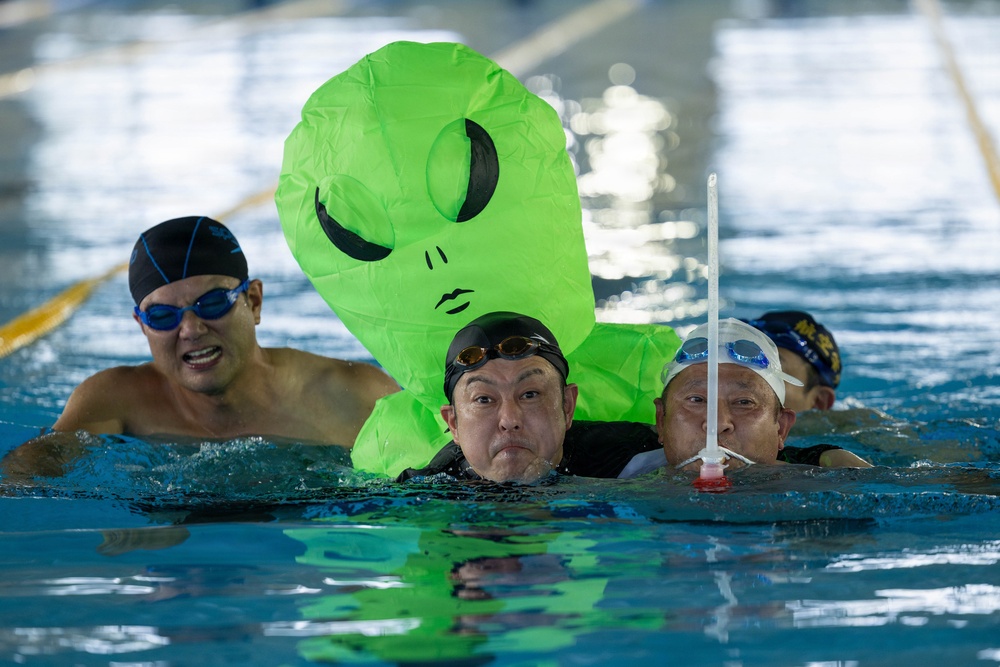 Several swimmers are in an indoor swimming pool, with one swimmer wearing a large inflatable green alien costume on their back. The scene appears to be a fun or novelty swimming event, possibly a race or recreational activity. The pool has lane markers visible in the background.
