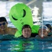Several swimmers are in an indoor swimming pool, with one swimmer wearing a large inflatable green alien costume on their back. The scene appears to be a fun or novelty swimming event, possibly a race or recreational activity. The pool has lane markers visible in the background.
