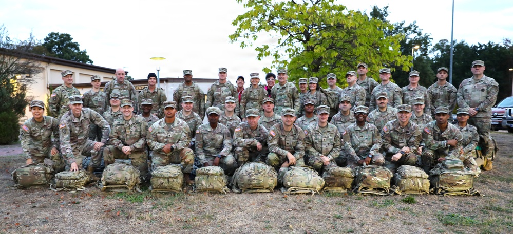 A large group of approximately 40 military personnel in U.S. Army OCP (Operational Camouflage Pattern) uniforms pose for a formal group photograph outdoors. The soldiers are arranged in three rows, with the front row kneeling behind a line of military rucksacks/ruck sacks placed on the ground. The setting appears to be a military installation, with a building visible in the background.