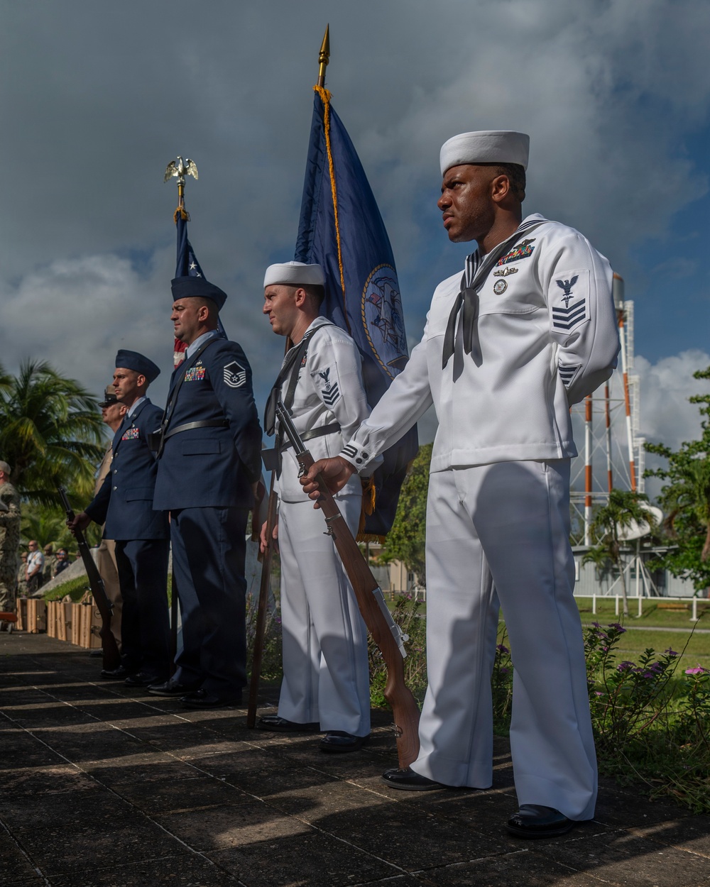 A joint military color guard ceremony is taking place outdoors, featuring personnel from the U.S. Navy and U.S. Air Force in their respective dress uniforms. Sailors in white dress uniforms carry rifles and a blue ceremonial flag, while Air Force personnel in service dress blues stand at attention. The setting appears to be a tropical location based on the palm trees and lush vegetation visible in the background.