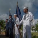 A joint military color guard ceremony is taking place outdoors, featuring personnel from the U.S. Navy and U.S. Air Force in their respective dress uniforms. Sailors in white dress uniforms carry rifles and a blue ceremonial flag, while Air Force personnel in service dress blues stand at attention. The setting appears to be a tropical location based on the palm trees and lush vegetation visible in the background.