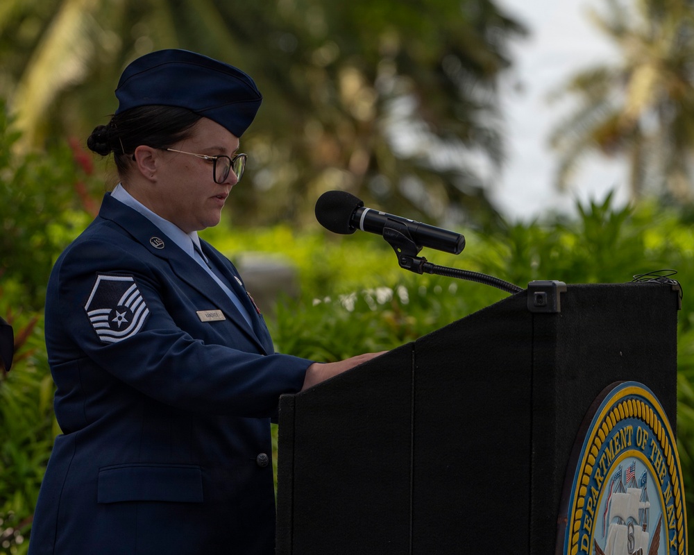A female U.S. Air Force Master Sergeant stands at a podium bearing the Department of the Navy seal, appearing to deliver remarks or read from notes at an official military ceremony. She is wearing the standard Air Force Service Dress uniform with a service cap, glasses, and visible rank insignia. The outdoor setting features tropical vegetation in the background, suggesting a location such as Guam, Hawaii, or another Pacific installation.
