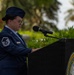 A female U.S. Air Force Master Sergeant stands at a podium bearing the Department of the Navy seal, appearing to deliver remarks or read from notes at an official military ceremony. She is wearing the standard Air Force Service Dress uniform with a service cap, glasses, and visible rank insignia. The outdoor setting features tropical vegetation in the background, suggesting a location such as Guam, Hawaii, or another Pacific installation.