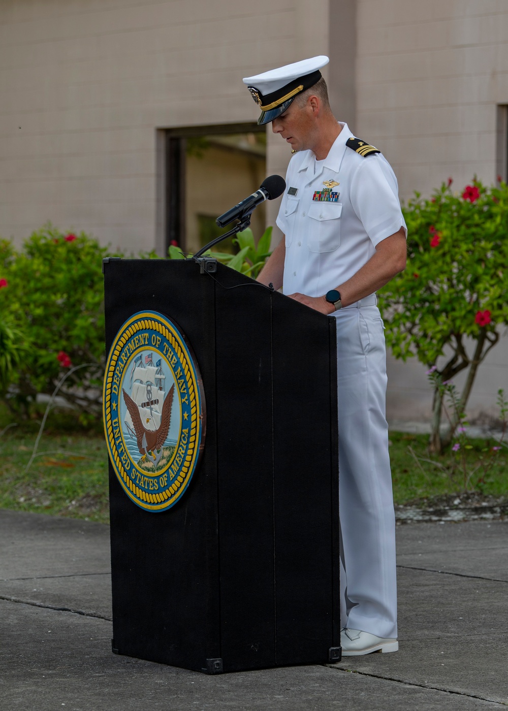 A U.S. Navy officer in white dress uniform stands at a podium bearing the official seal of the Department of the Navy, United States of America. The officer appears to be delivering a speech or remarks at an outdoor military ceremony. The setting includes a building and tropical vegetation in the background, suggesting a location in a warm climate.
