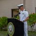 A U.S. Navy officer in white dress uniform stands at a podium bearing the official seal of the Department of the Navy, United States of America. The officer appears to be delivering a speech or remarks at an outdoor military ceremony. The setting includes a building and tropical vegetation in the background, suggesting a location in a warm climate.