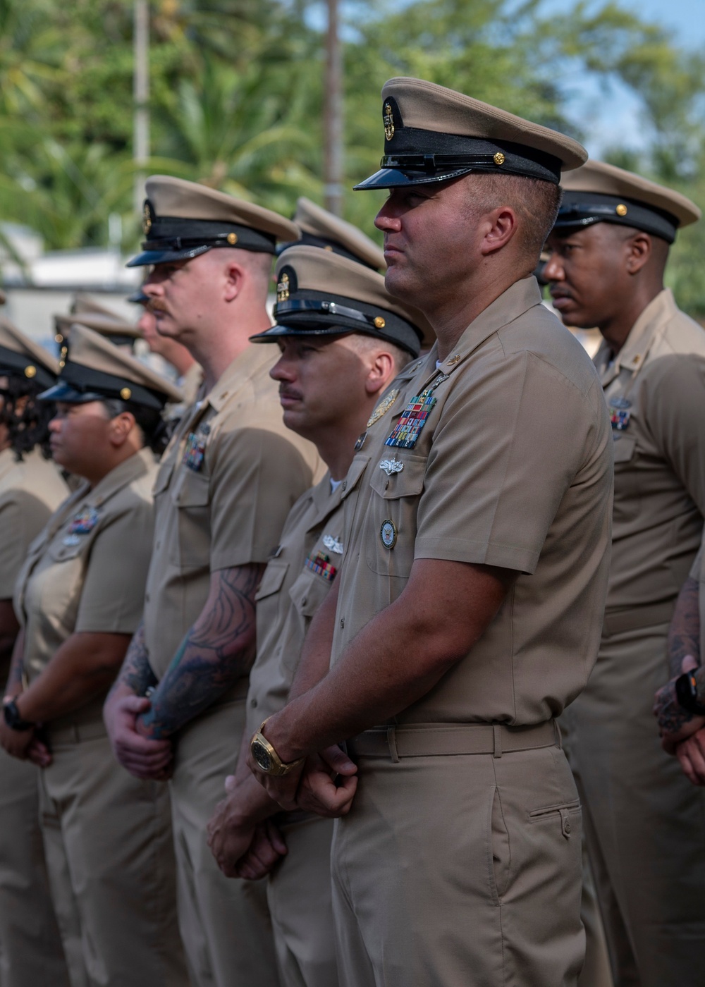 A group of United States Navy Chief Petty Officers standing in formation at what appears to be an outdoor military ceremony. The personnel are wearing the Navy Service Uniform (NSU) in tan/khaki with combination covers (peaked caps). The setting appears to be in a tropical location based on the palm trees visible in the background.
