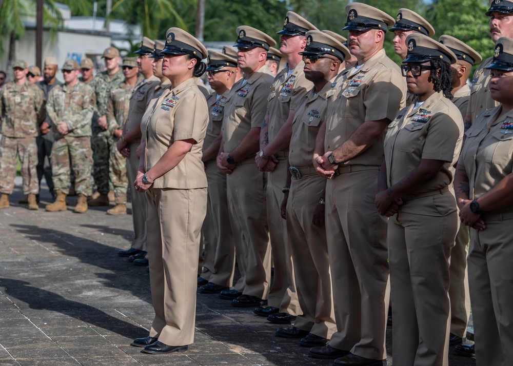 A formal military ceremony featuring U.S. Navy personnel in khaki service uniforms standing at attention in organized formation. In the background, additional military personnel in Army combat uniforms (ACUs) are also present, suggesting a joint-service event. The setting appears to be an outdoor parade ground or courtyard, likely at a military installation in a tropical location.