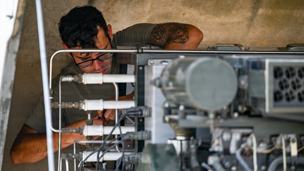 A technician wearing glasses is closely inspecting or working on a complex mechanical or hydraulic system with multiple pipes, fittings, and connectors. The individual has a visible tattoo on their upper arm and is wearing an olive/military-green t-shirt. The setting appears to be an industrial or military maintenance environment.