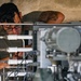 A technician wearing glasses is closely inspecting or working on a complex mechanical or hydraulic system with multiple pipes, fittings, and connectors. The individual has a visible tattoo on their upper arm and is wearing an olive/military-green t-shirt. The setting appears to be an industrial or military maintenance environment.