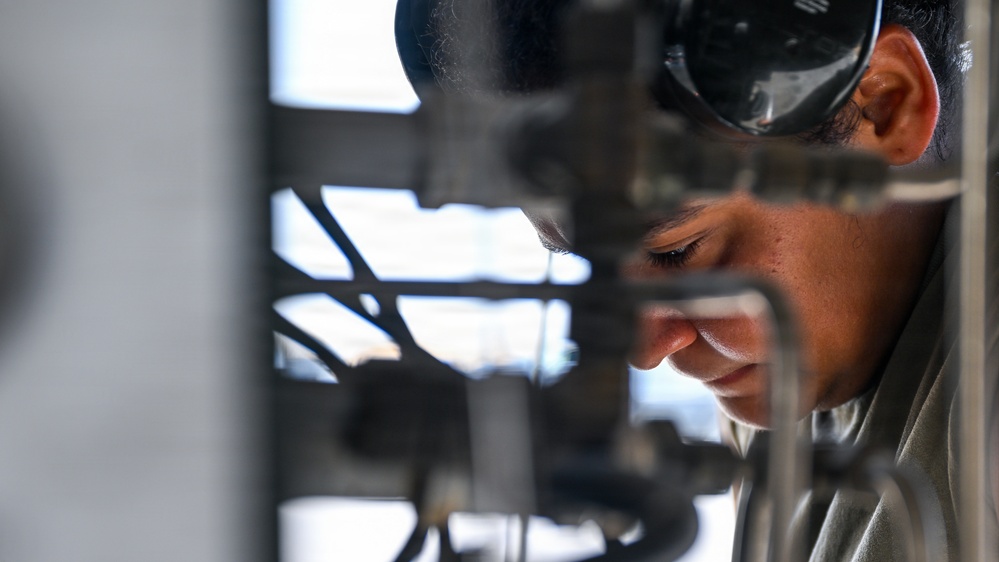 A person wearing protective goggles pushed up on their forehead is closely inspecting or working on mechanical equipment. The individual appears focused, leaning in to examine pipes, tubes, or structural components in what appears to be an industrial or maintenance setting. The shallow depth of field emphasizes the worker's face against a blurred background of metal framework.
