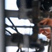 A person wearing protective goggles pushed up on their forehead is closely inspecting or working on mechanical equipment. The individual appears focused, leaning in to examine pipes, tubes, or structural components in what appears to be an industrial or maintenance setting. The shallow depth of field emphasizes the worker's face against a blurred background of metal framework.
