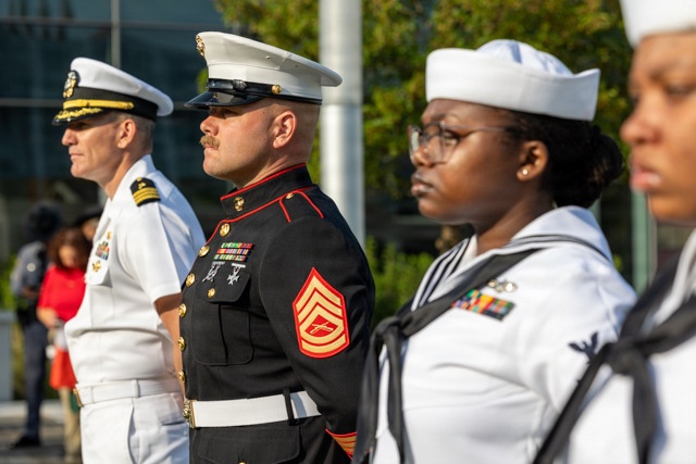 The image shows a formal military ceremony featuring personnel from multiple U.S. armed service branches standing at attention. Visible are a U.S. Navy officer in dress whites, a U.S. Marine Corps Staff Non-Commissioned Officer in dress blues, and two U.S. Navy enlisted sailors in their dress white uniforms. The setting appears to be an outdoor ceremonial event, likely a parade, memorial, or official military observance.