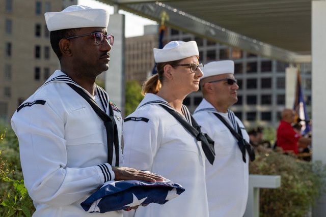 Three U.S. Navy sailors in dress white uniforms stand at attention during what appears to be a formal military ceremony. The sailor in the foreground holds a folded American flag, suggesting this may be a memorial or commemorative event. The setting appears to be an outdoor venue with urban buildings visible in the background.