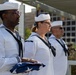 Three U.S. Navy sailors in dress white uniforms stand at attention during what appears to be a formal military ceremony. The sailor in the foreground holds a folded American flag, suggesting this may be a memorial or commemorative event. The setting appears to be an outdoor venue with urban buildings visible in the background.
