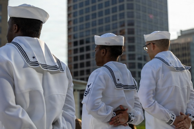 Three U.S. Navy sailors in dress white uniforms are standing at parade rest in an outdoor urban setting. They are viewed primarily from behind, with one sailor partially visible in profile. A modern high-rise building is visible in the background.