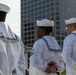 Three U.S. Navy sailors in dress white uniforms are standing at parade rest in an outdoor urban setting. They are viewed primarily from behind, with one sailor partially visible in profile. A modern high-rise building is visible in the background.