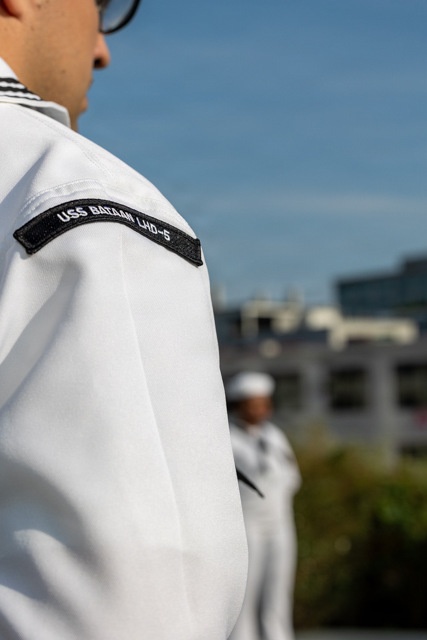 A U.S. Navy sailor in white dress uniform is shown from behind/side, wearing a shoulder patch identifying their assignment to USS Bataan LHD-5, an amphibious assault ship. At least one additional sailor in white uniform is visible in the blurred background. The setting appears to be an outdoor formation or ceremony on a clear, sunny day.