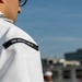 A U.S. Navy sailor in white dress uniform is shown from behind/side, wearing a shoulder patch identifying their assignment to USS Bataan LHD-5, an amphibious assault ship. At least one additional sailor in white uniform is visible in the blurred background. The setting appears to be an outdoor formation or ceremony on a clear, sunny day.