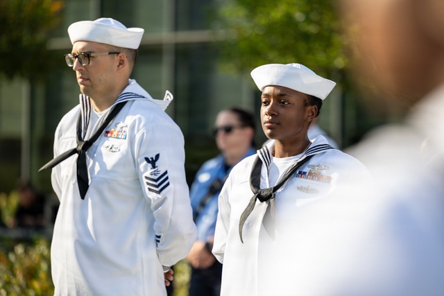 Two U.S. Navy sailors in dress white uniforms stand at attention during what appears to be a formal military ceremony or event. The sailor on the left displays Petty Officer rating chevrons and multiple service ribbons, while the sailor on the right also wears ribbons and a qualification badge. Both wear the traditional white Dixie cup covers and black neckerchiefs.