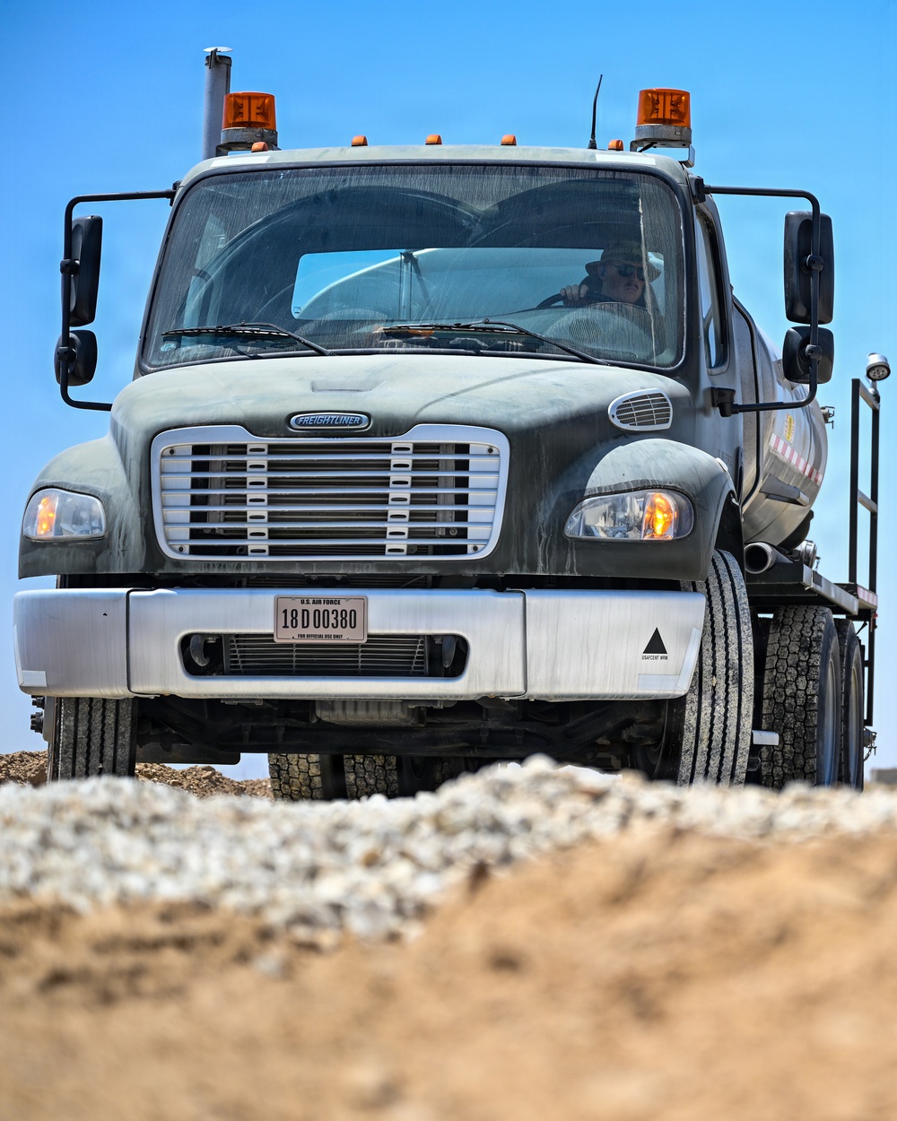 A U.S. Air Force Freightliner heavy-duty truck is photographed head-on from a low angle on what appears to be a construction or unpaved work site. The vehicle bears an official U.S. Air Force license plate reading '18D00380 - FOR OFFICIAL USE ONLY' and is equipped with amber warning lights on the roof. A driver wearing sunglasses is visible through the windshield.