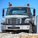 A U.S. Air Force Freightliner heavy-duty truck is photographed head-on from a low angle on what appears to be a construction or unpaved work site. The vehicle bears an official U.S. Air Force license plate reading '18D00380 - FOR OFFICIAL USE ONLY' and is equipped with amber warning lights on the roof. A driver wearing sunglasses is visible through the windshield.