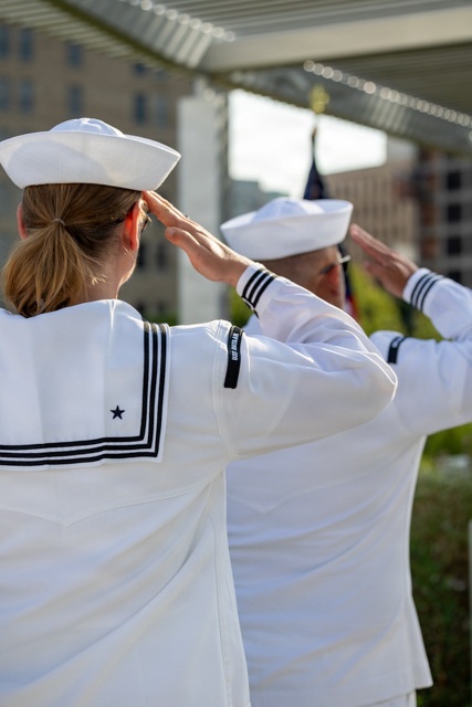 Two U.S. Navy sailors in white dress uniforms (Dress Whites) are photographed from behind while rendering a salute. Both wear the distinctive white 'Dixie cup' sailor caps and traditional jumper-style uniforms with black piping and stripes on the sleeves. A flag is visible in the blurred background, suggesting a formal military ceremony.