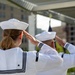 Two U.S. Navy sailors in white dress uniforms (Dress Whites) are photographed from behind while rendering a salute. Both wear the distinctive white 'Dixie cup' sailor caps and traditional jumper-style uniforms with black piping and stripes on the sleeves. A flag is visible in the blurred background, suggesting a formal military ceremony.