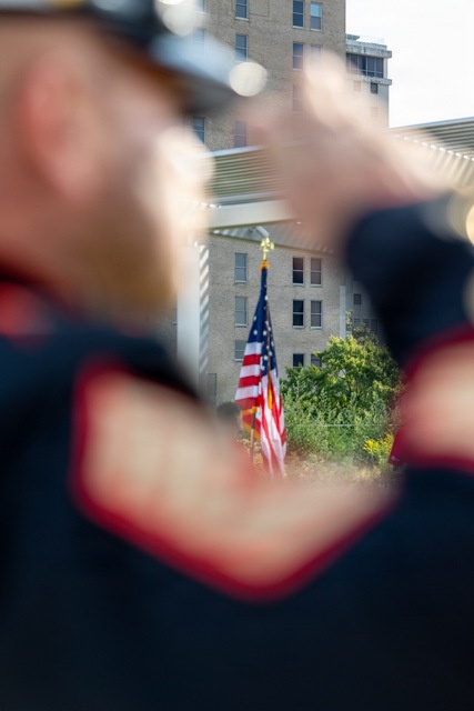 A close-up, shallow depth-of-field photograph taken from behind a uniformed individual who appears to be performing a military or ceremonial rifle salute. In the sharp background, an American flag on a pole stands in front of a multi-story brick building surrounded by green foliage. The foreground is intentionally blurred, framing the flag through the gap between the person's arms and rifle.