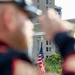 A close-up, shallow depth-of-field photograph taken from behind a uniformed individual who appears to be performing a military or ceremonial rifle salute. In the sharp background, an American flag on a pole stands in front of a multi-story brick building surrounded by green foliage. The foreground is intentionally blurred, framing the flag through the gap between the person's arms and rifle.