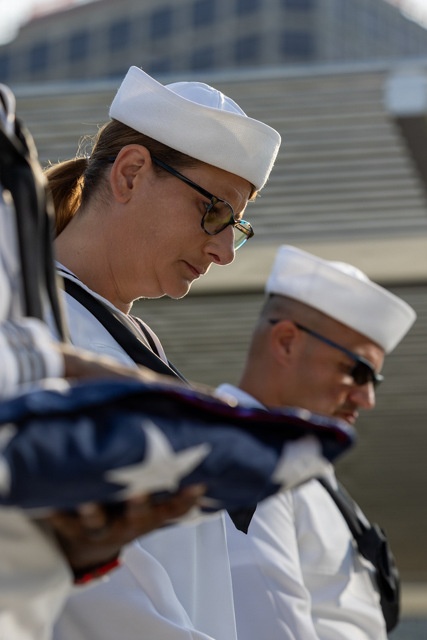 Two U.S. Navy sailors in dress white uniforms are participating in a formal military ceremony, with heads bowed in a solemn posture. A folded American flag is visible in the foreground, indicating this is likely a funeral honors or memorial service. The image captures a moment of reverence and military protocol.