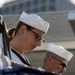 Two U.S. Navy sailors in dress white uniforms are participating in a formal military ceremony, with heads bowed in a solemn posture. A folded American flag is visible in the foreground, indicating this is likely a funeral honors or memorial service. The image captures a moment of reverence and military protocol.