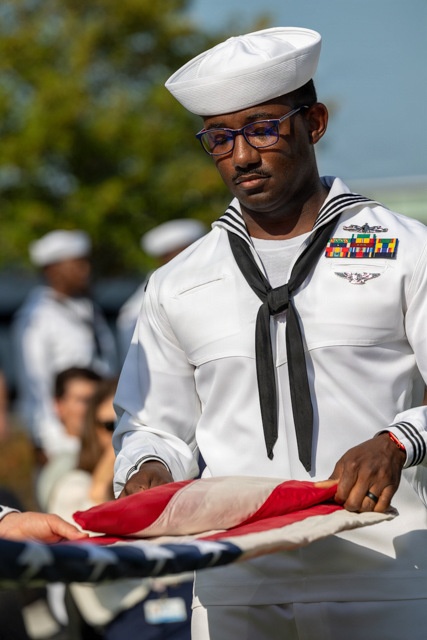 A U.S. Navy sailor in dress white uniform is carefully folding or handling an American flag, likely during a military ceremony or funeral honors detail. The sailor wears multiple service ribbons, a warfare qualification badge, and the traditional white dixie cup cover with black neckerchief. Other sailors in white uniforms are visible in the blurred background alongside civilian observers.