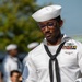 A U.S. Navy sailor in dress white uniform is carefully folding or handling an American flag, likely during a military ceremony or funeral honors detail. The sailor wears multiple service ribbons, a warfare qualification badge, and the traditional white dixie cup cover with black neckerchief. Other sailors in white uniforms are visible in the blurred background alongside civilian observers.