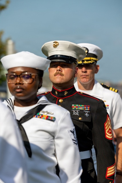 Three U.S. military personnel in dress uniforms are standing in formation, likely during a formal ceremony or parade. The group includes a U.S. Navy enlisted sailor (foreground), a U.S. Marine Corps Staff Non-Commissioned Officer in dress blues (center), and a U.S. Navy officer (background). All individuals are wearing service medals and ribbons, indicating experienced service members.