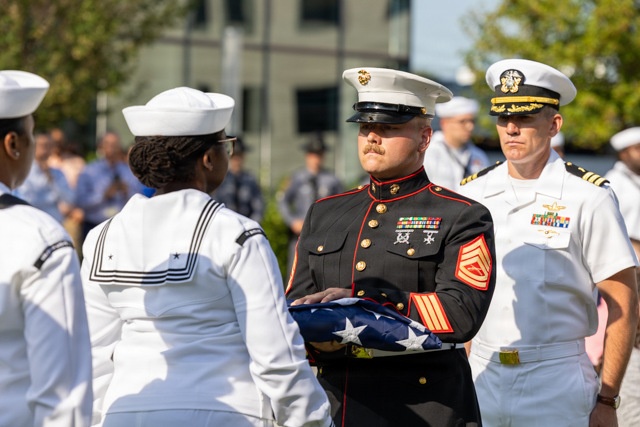 A formal military ceremony is taking place outdoors, featuring personnel from the United States Marine Corps and United States Navy in dress uniforms. A Marine Staff Sergeant in dress blues is holding a folded American flag, likely during a memorial or honors ceremony. A Navy officer with command insignia and ribbons observes in the background alongside multiple Navy sailors in white dress uniforms.