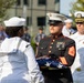 A formal military ceremony is taking place outdoors, featuring personnel from the United States Marine Corps and United States Navy in dress uniforms. A Marine Staff Sergeant in dress blues is holding a folded American flag, likely during a memorial or honors ceremony. A Navy officer with command insignia and ribbons observes in the background alongside multiple Navy sailors in white dress uniforms.