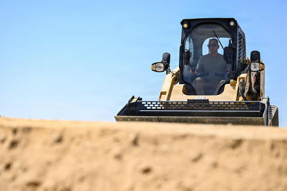 A person is operating a Caterpillar (CAT) compact track loader (skid steer) on a sandy construction or earthmoving site. The machine is yellow and black, consistent with CAT branding, and is positioned atop or near a mound of sand. The image is taken from a low angle, emphasizing the scale of the machine against a clear blue sky.