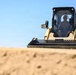 A person is operating a Caterpillar (CAT) compact track loader (skid steer) on a sandy construction or earthmoving site. The machine is yellow and black, consistent with CAT branding, and is positioned atop or near a mound of sand. The image is taken from a low angle, emphasizing the scale of the machine against a clear blue sky.