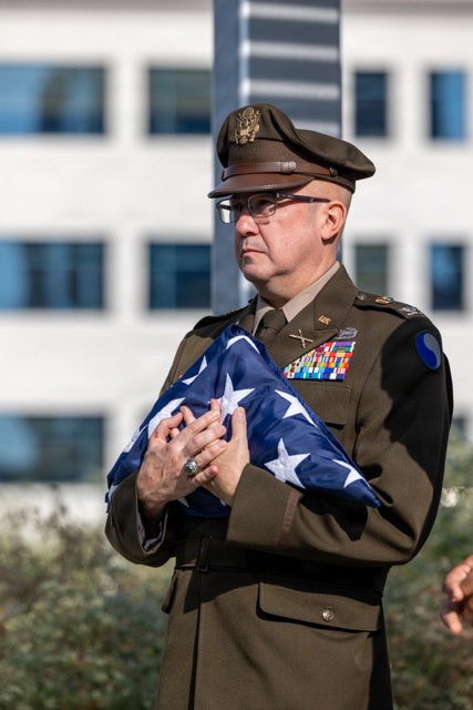 A U.S. Army officer in dress uniform (Army Greens) holds a ceremonially folded American flag against his chest during what appears to be a formal military ceremony. The officer displays an extensive array of service ribbons and medals on his chest, along with branch insignia and a shoulder patch. The setting appears to be outdoors near a modern office or government building.