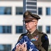 A U.S. Army officer in dress uniform (Army Greens) holds a ceremonially folded American flag against his chest during what appears to be a formal military ceremony. The officer displays an extensive array of service ribbons and medals on his chest, along with branch insignia and a shoulder patch. The setting appears to be outdoors near a modern office or government building.