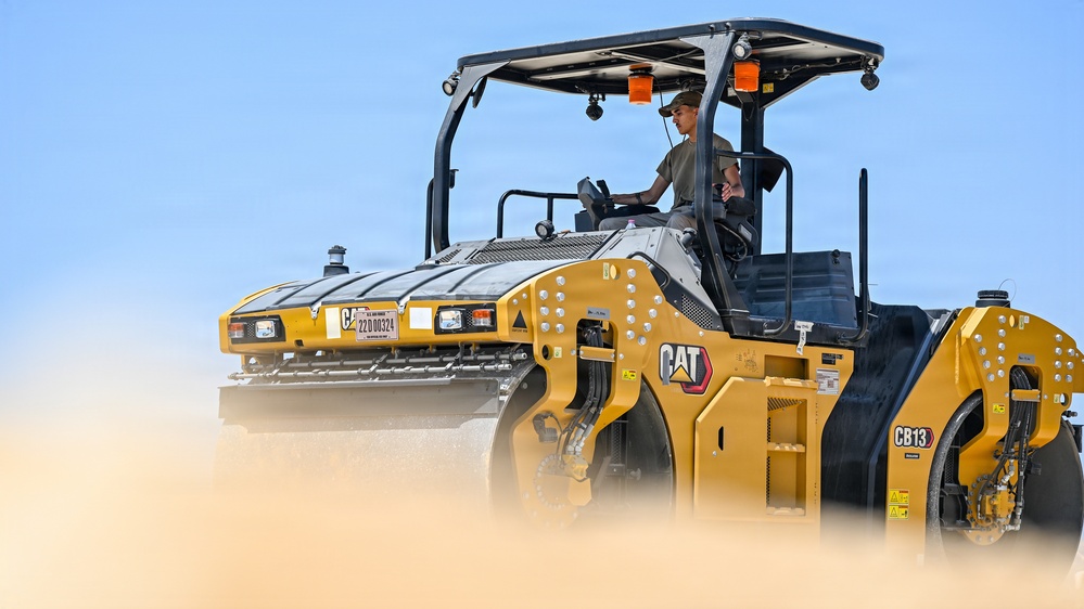 A Caterpillar CB13 tandem vibratory asphalt compactor roller is being operated by a single worker on what appears to be a construction or road-building site. The machine is yellow and black, consistent with CAT branding, and is generating dust or steam as it operates. The image is a professional, high-quality photograph taken from a low angle against a clear blue sky.