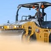 A Caterpillar CB13 tandem vibratory asphalt compactor roller is being operated by a single worker on what appears to be a construction or road-building site. The machine is yellow and black, consistent with CAT branding, and is generating dust or steam as it operates. The image is a professional, high-quality photograph taken from a low angle against a clear blue sky.