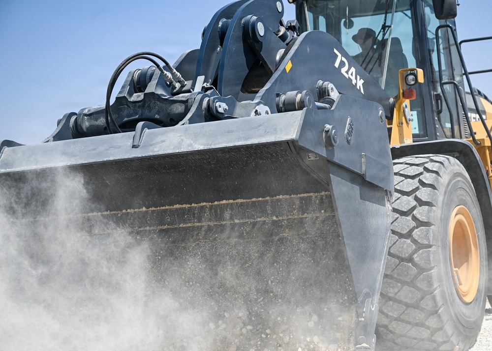 A close-up photograph of a John Deere 724K wheel loader actively scooping material, with visible dust being kicked up from the ground. The machine is yellow and black, photographed from a low angle emphasizing the large bucket and front tire. An operator is visible inside the cab in the background.