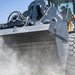 A close-up photograph of a John Deere 724K wheel loader actively scooping material, with visible dust being kicked up from the ground. The machine is yellow and black, photographed from a low angle emphasizing the large bucket and front tire. An operator is visible inside the cab in the background.