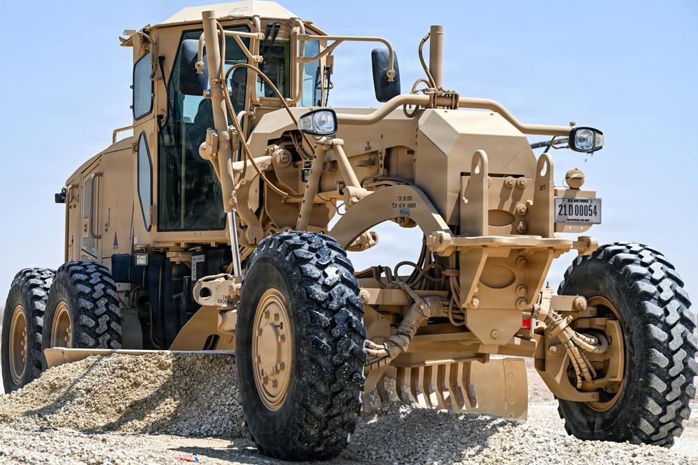 A U.S. Air Force motor grader painted in desert tan/sand color is photographed at close range on a gravel or sandy terrain. The vehicle bears a U.S. Air Force registration plate reading '21D 00054'. It appears to be engaged in road grading or construction operations in an arid environment.